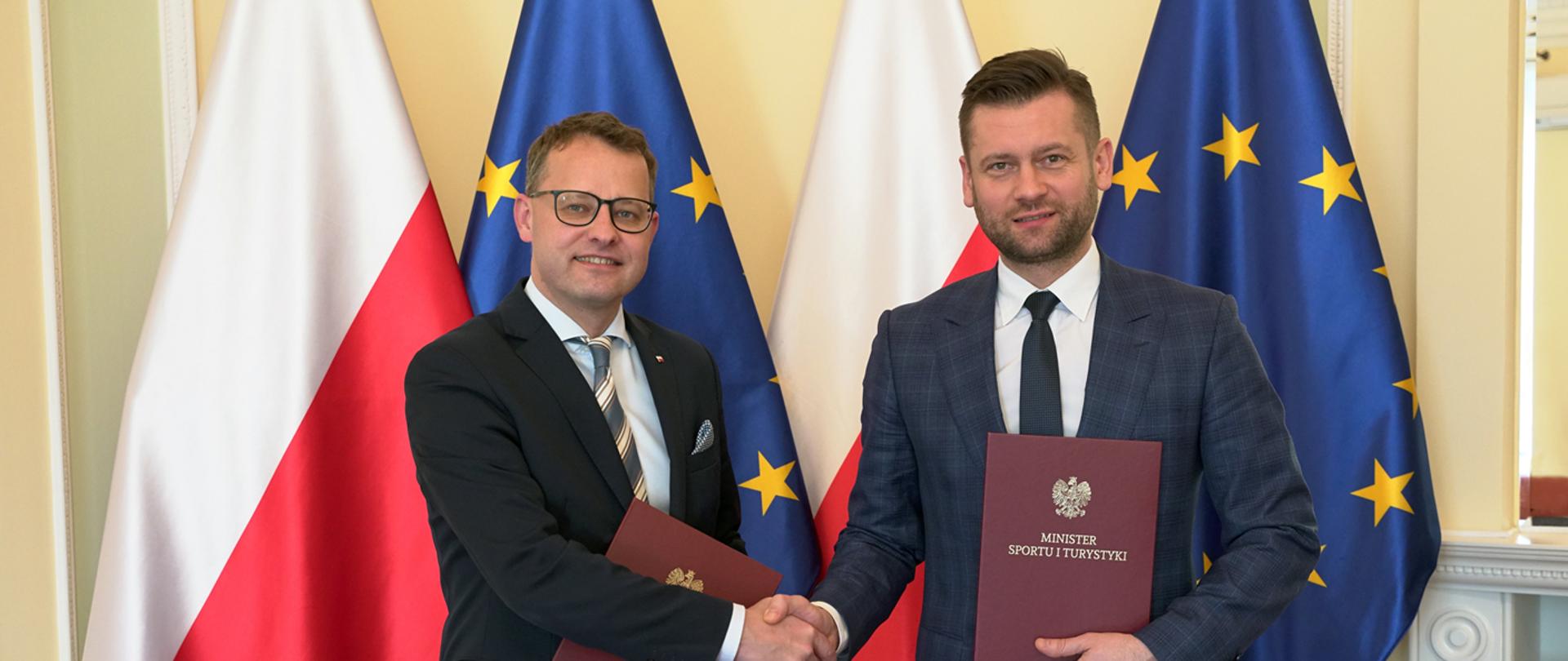 Porozumienie w sprawie pomocy sportowcom i zapobieganiu przestępczości w sporcie; minister sportu i turystyki Kamil Bortniczuk i podsekretarz stanu w ministerstwie sprawiedliwości Marcin Romanowski przekazują sobie uścisk dłoni trzymając podpisane porozumienie, na tle flag Polski i UE