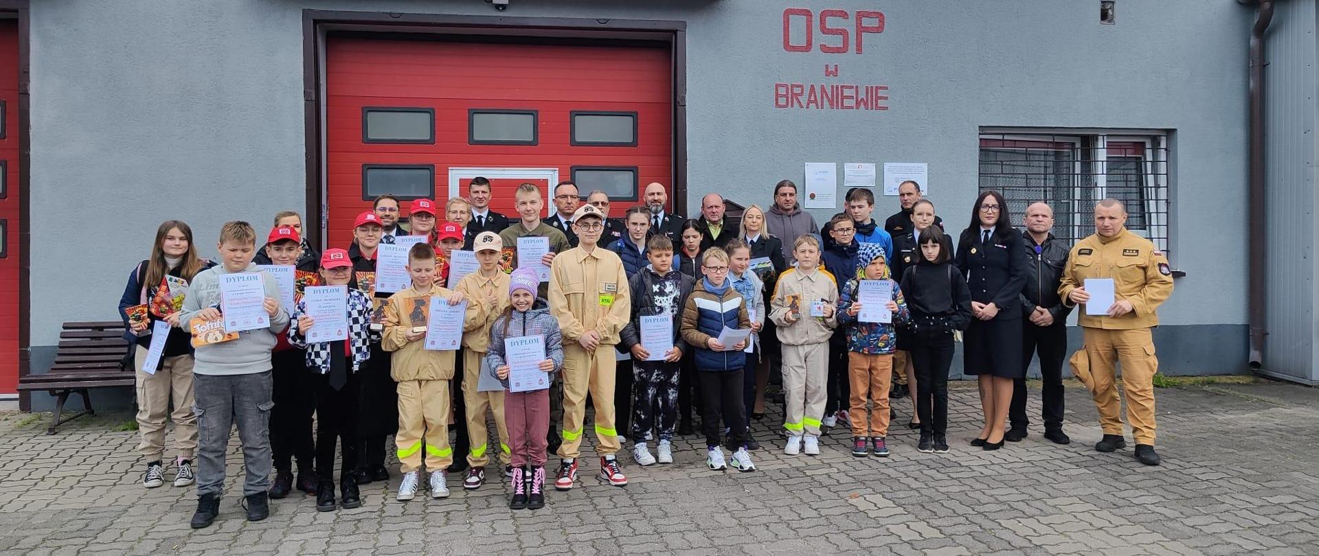 Na zdjęciu na tle remizy OSP Braniewo stoi duża grupa dzieci z dyplomami. W śród dzieci organizatorzy i wychowawcy dzieci ze szkół.