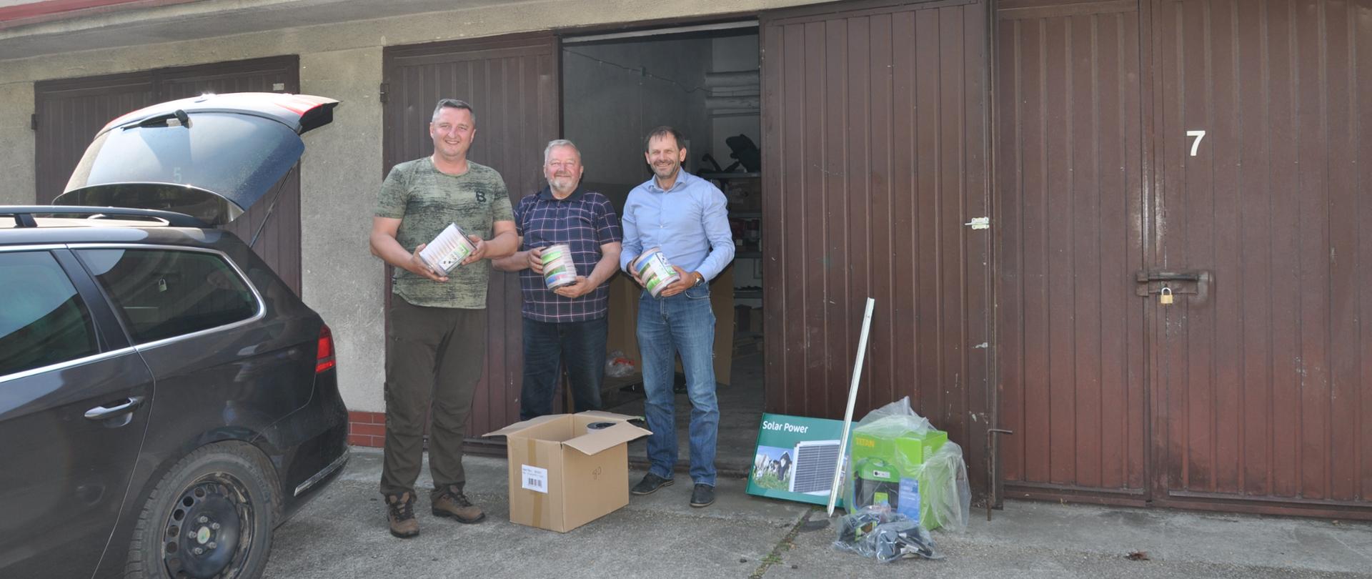 Trzech mężczyzn pozuje do zdjęcia, w rękach trzymają szpule ze zwiniętym drutem, na ziemi karton oraz kilka urządzeń. Za ich plecami brązowe drzwi od garażu.