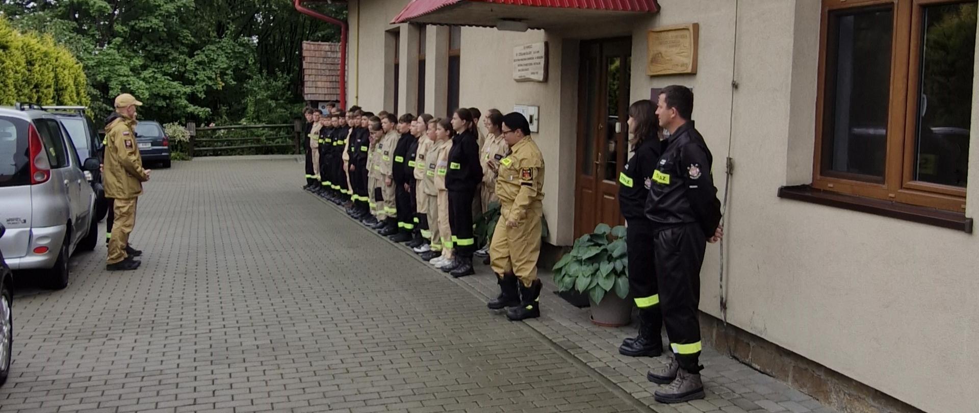Grupa strażaków jest ustawiona w szeregu na zewnątrz jasnego budynku, z czerwonym dachem. Stoją wzdłuż ściany po prawej stronie, ubrani w różne wersje mundurów strażackich - zarówno w jasne, jak i ciemne kombinezony. Po lewej stronie, wzdłuż linii zaparkowanych aut stoi strażak w jasnym koszarze, zwrócony jest twarzą do pozostałych strażaków i wygląda jakby przemawiał do nich lub wydawał polecenia.