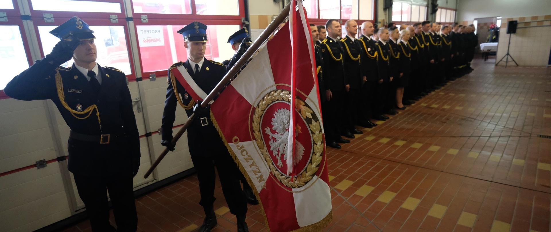 Poczet sztandarowy KP PSP Chojnice oraz pododdział strażaków na tle bram garażowych.