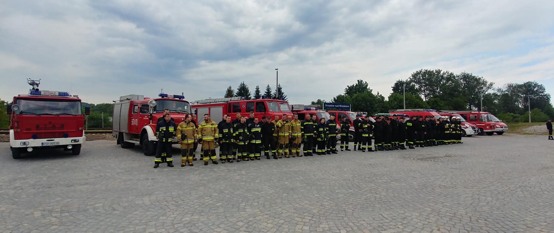 Na zdjęciu widoczna zbiórka pododdziałów jednostek OSP z terenu Powiatu Strzyżowskiego. Za nimi widoczne samochody pożarnicze. W tle drzewa