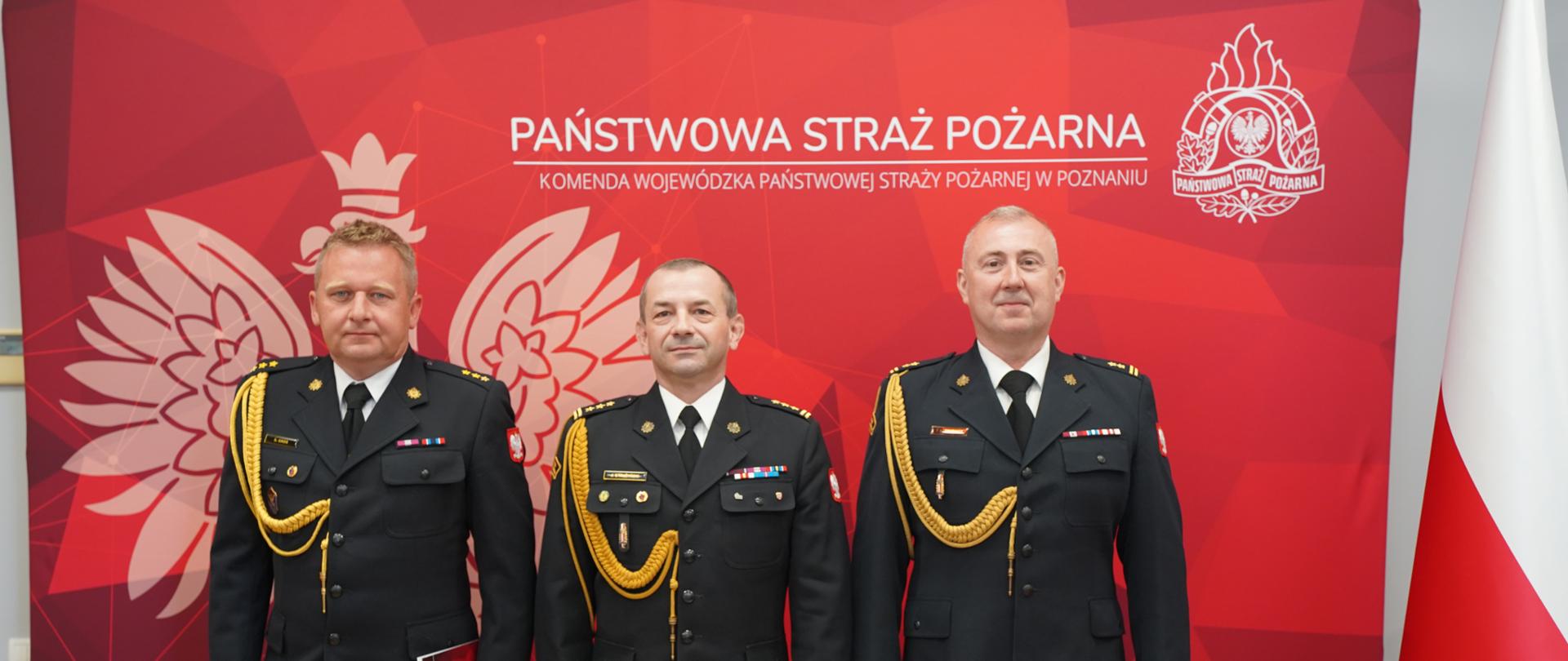 Trzech strażaków Państwowej Straży Pożarnej w galowych mundurach pozuje do oficjalnego zdjęcia na tle czerwonej ścianki Komendy Wojewódzkiej PSP w Poznaniu. Obok widoczna jest flaga Polski oraz emblemat straży pożarnej.