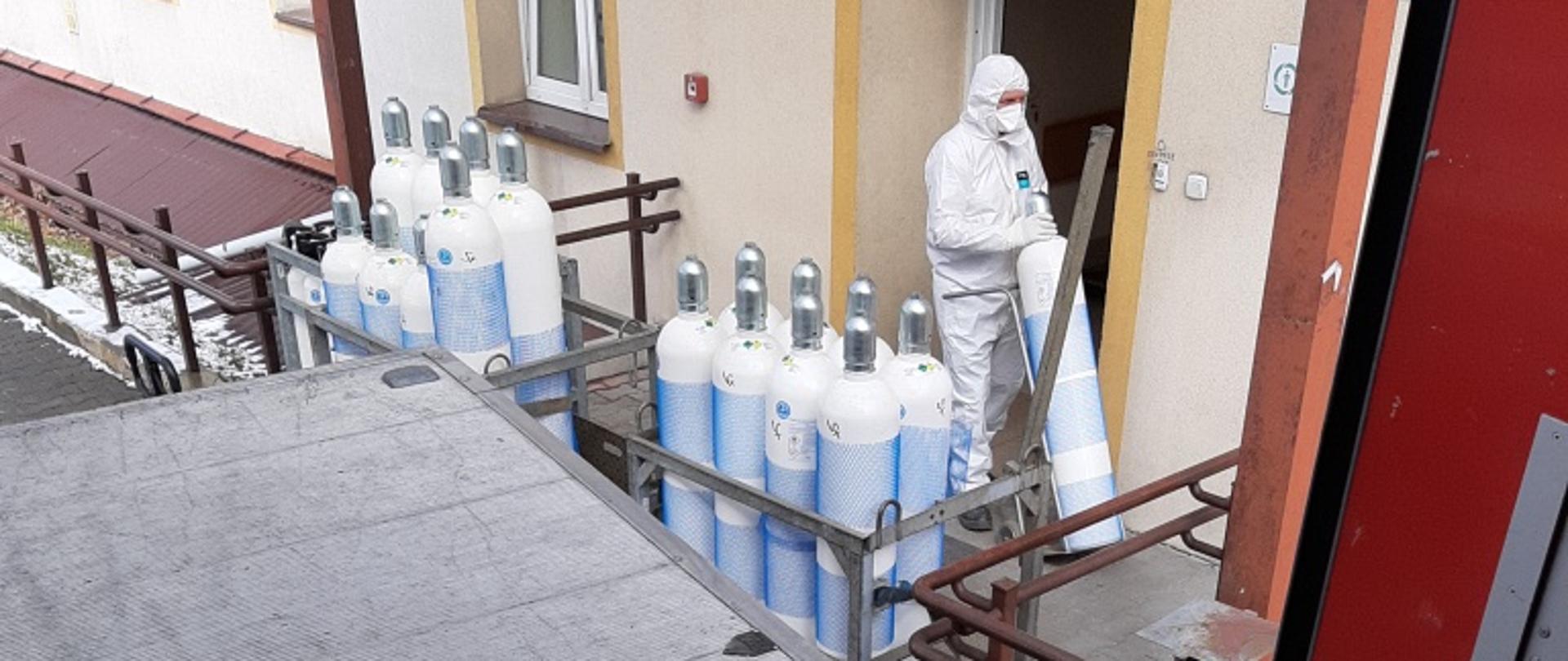 Zdjęcie wykonane z przestrzeni transportowej pojazdu ciężarowego. Na zdjęciu widać pracownika szpitala ubranego w specjalną odzież ochronną. Pracownik wnosi butle dostarczone pod szpital do pomieszczeń zlokalizowanych wewnątrz budynku. 