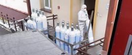 Zdjęcie wykonane z przestrzeni transportowej pojazdu ciężarowego. Na zdjęciu widać pracownika szpitala ubranego w specjalną odzież ochronną. Pracownik wnosi butle dostarczone pod szpital do pomieszczeń zlokalizowanych wewnątrz budynku. 