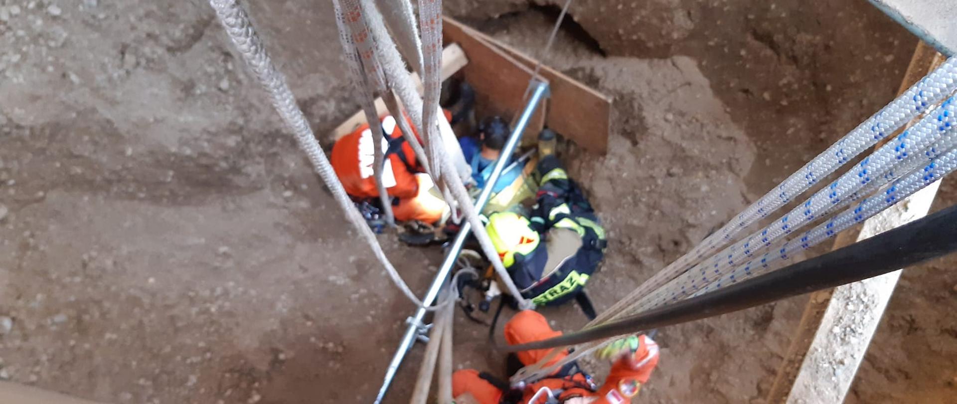 Widok z góry na ratowników Specjalistycznej Grupy Poszukiwawczo Ratowniczej Gdańsk stabilizujących grunt wokół poszkodowanego w zbiorniku ze żwirem, a także na liny służące do asekuracji i opuszczania ratowników.