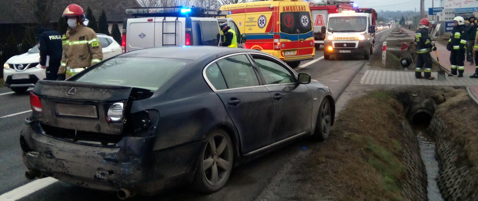 Na pierwszym planie znajduje się samochód osobowy marki Lexus z uszkodzonym tylnym zderzakiem. W tle znajduje się strażak oraz samochody Policji, Pogotowia Ratunkowego i Straży Pożarnej. Po prawej stronie znajdują się strażacy porządkujący teren akcji.