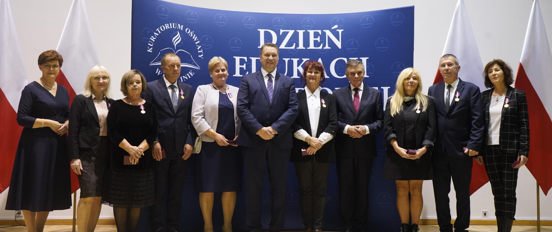 Pod scianą na której wisi niebieski baner z napisem Dzień edukacji stoi grupa ludzi, za nimi po bokach polskie flagi.