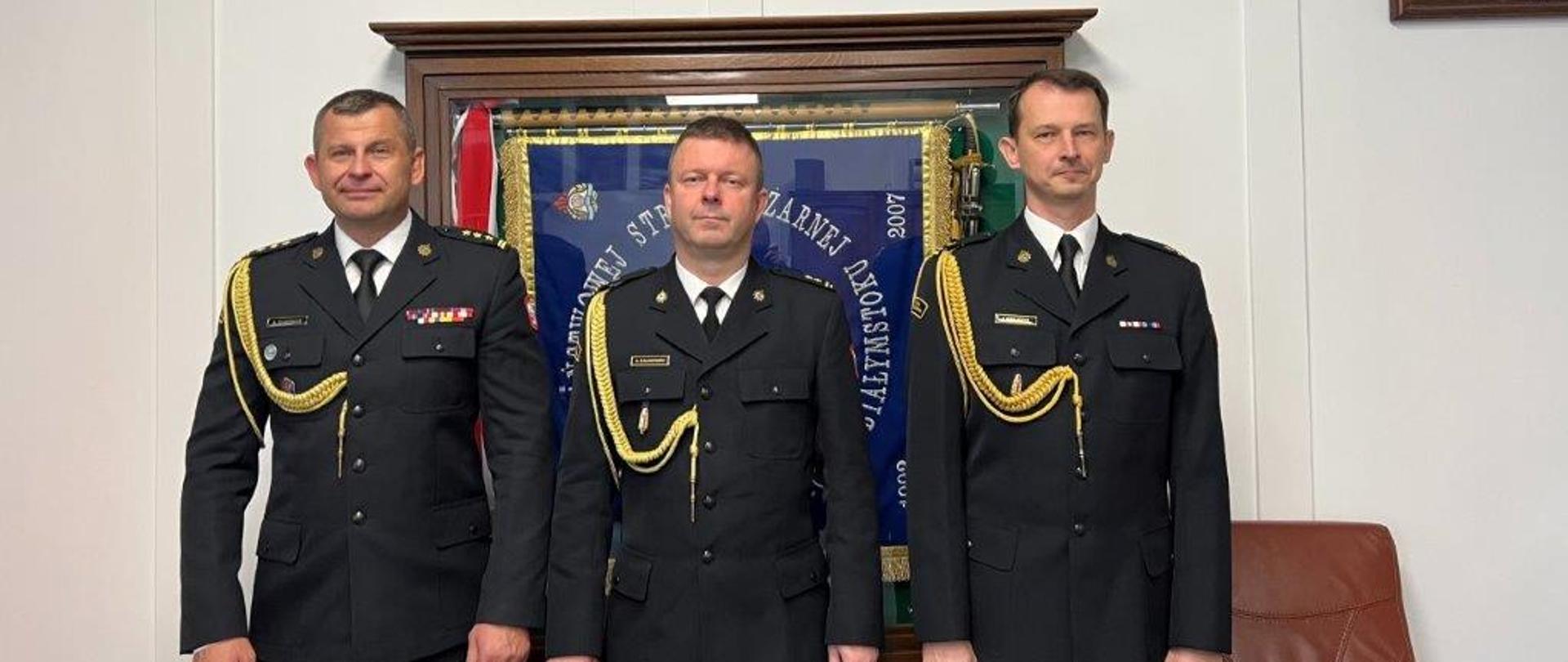 4. Trzech Funkcjonariuszy Państwowej Straży Pożarnej ubranych w mundury wyjściowe pozuje do zdjęcia. W tle widoczny jest sztandar Komendy Wojewódzkiej Państwowej Straży Pożarnej w Białymstoku schowany w przeszklonej gablocie. 