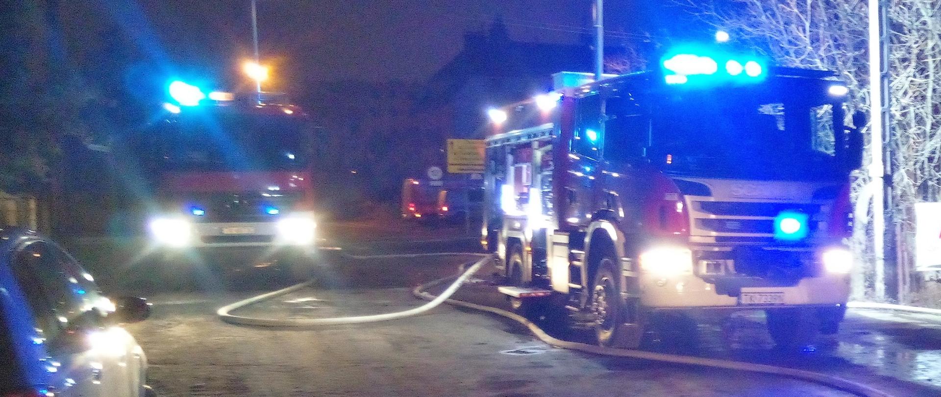 Zdjęcie przedstawia ulicę przy której doszło do pożaru domu. Zaparkowały na niej trzy samochody pożarnicze z których rozciągnięto węże gaśnicze.