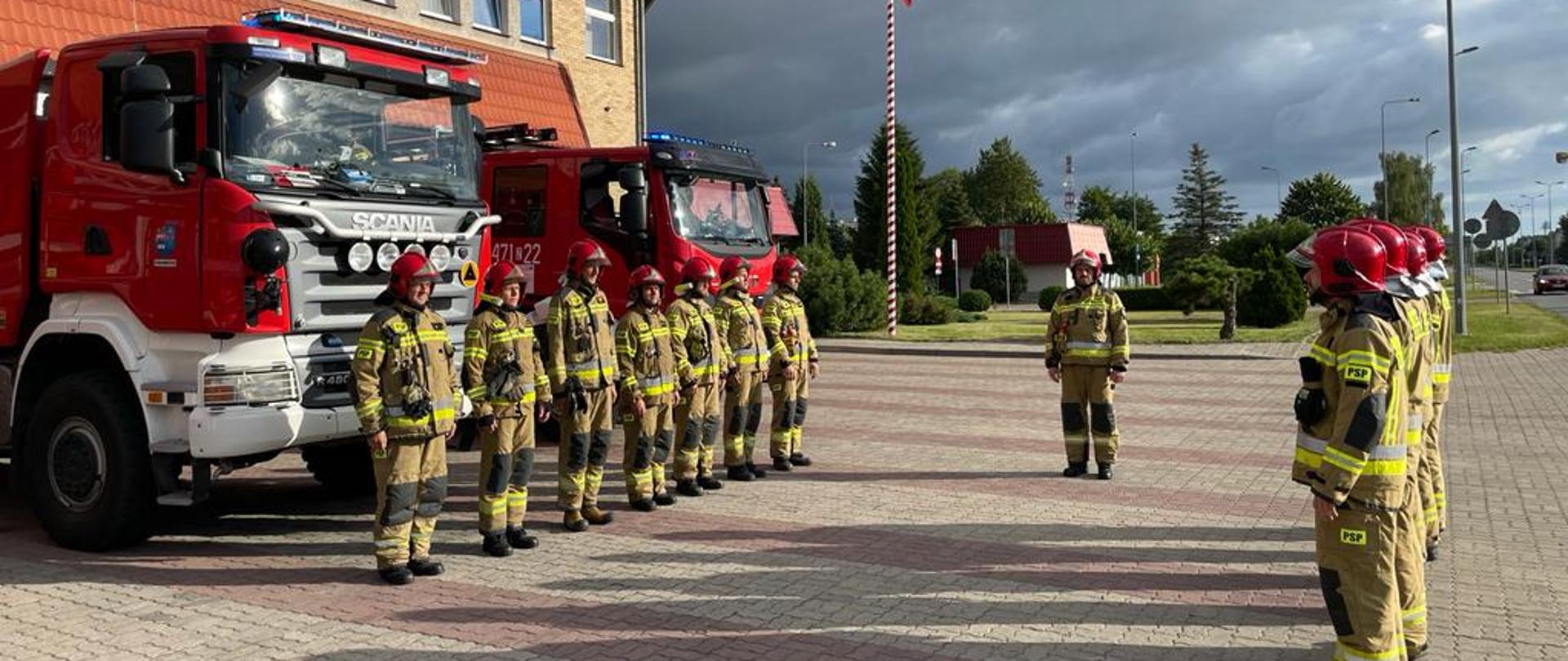 Strażacy stojący w dwóch rzędach oddają hołd tragicznie zmarłemu strażakowi, w tle czerwone samochody strażackie.