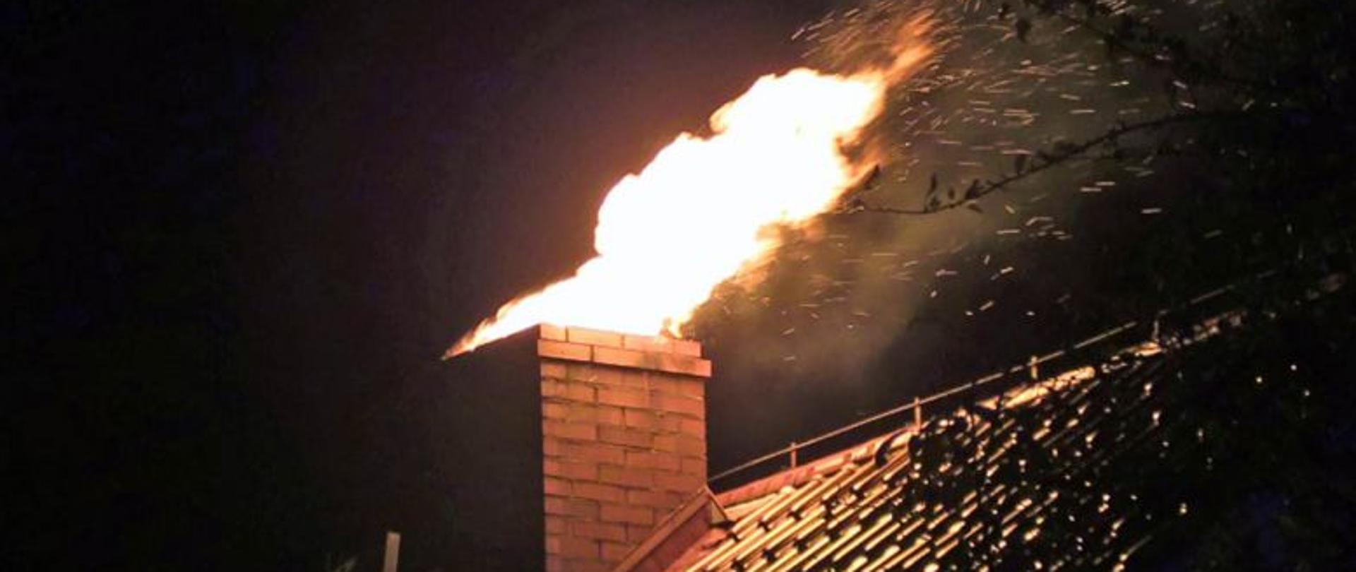 Na zdjęciu przedstawiony jest dach budynku wraz z kominem, z którego wydobywa się ogień oraz iskry. Zdjęcie zrobione w nocy, dookoła panuje mrok.