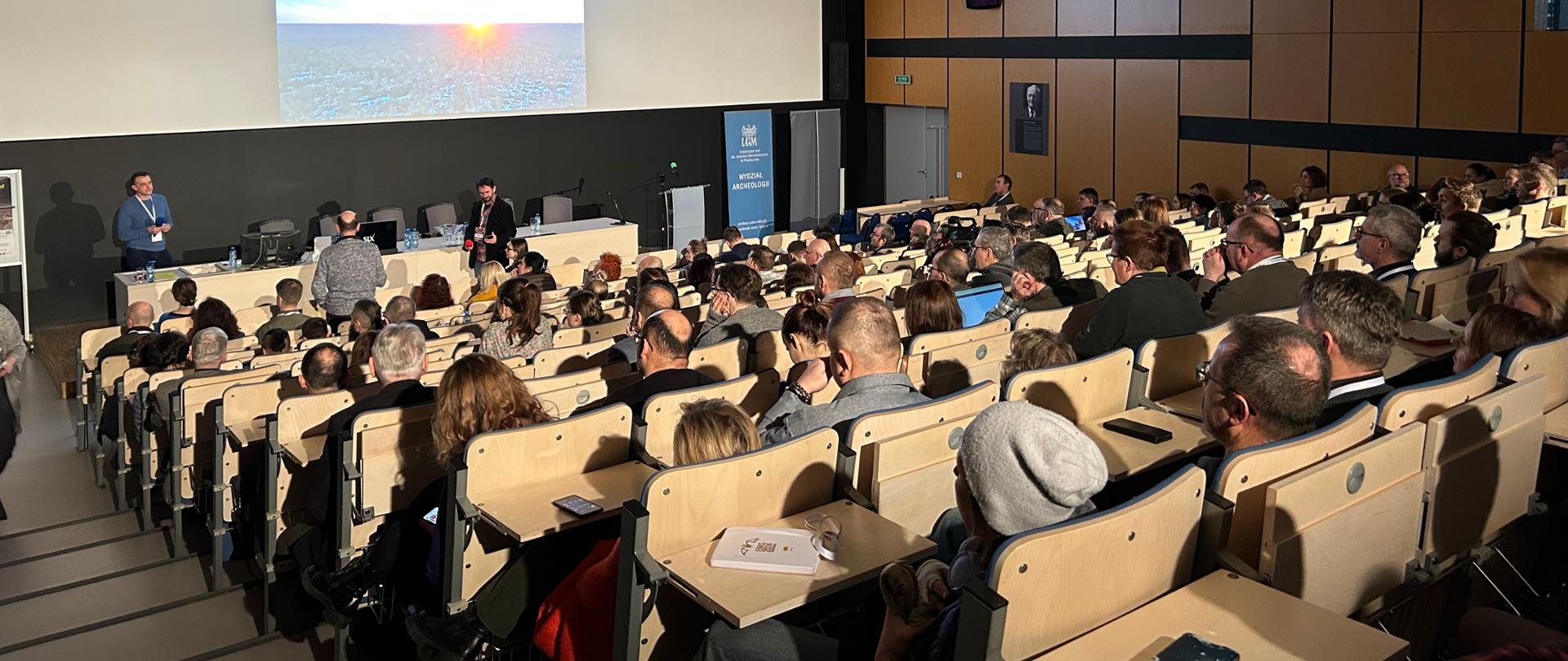 Duża aula. Ludzie siedzą rzędami. Z przodu mężczyzna trzyma mikrofon i przemawia do zebranych. Na ekranie za jego plecami wyświetlana jest prezentacja. 