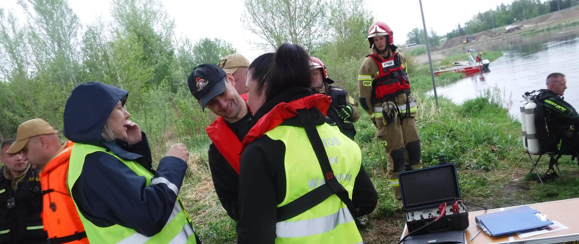 Inspektorzy WIOŚ w Warszawie rozmawiają ze strażakiem nad brzegiem rzeki. W tle przemieszcza się grupa strażaków w pełnym umundurowaniu, a na brzegu rzeki siedzi nurek.