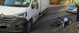 Motorower leży na jezdni, obok stoi biały samochód transportowy marki Renault. Po prawo samochód policyjny.