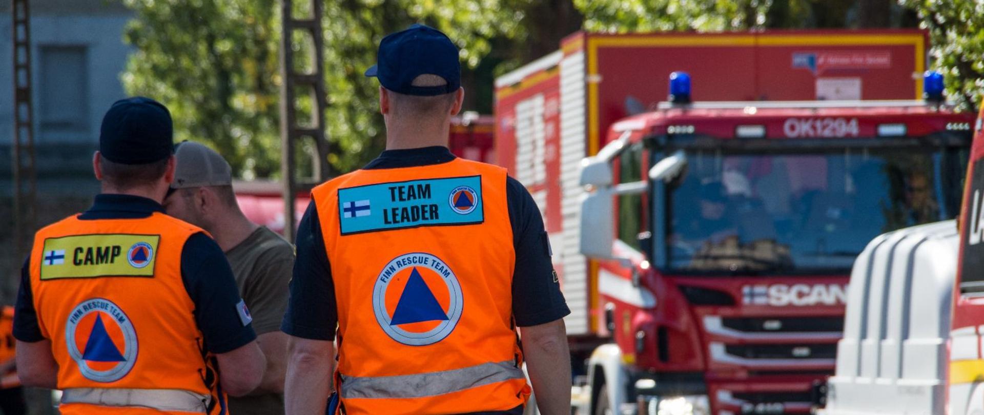 Dwaj mężczyźni z kamizelkami z logiem obrony cywilnej przyglądający się wjeżdzającej kolumnie pojazdów