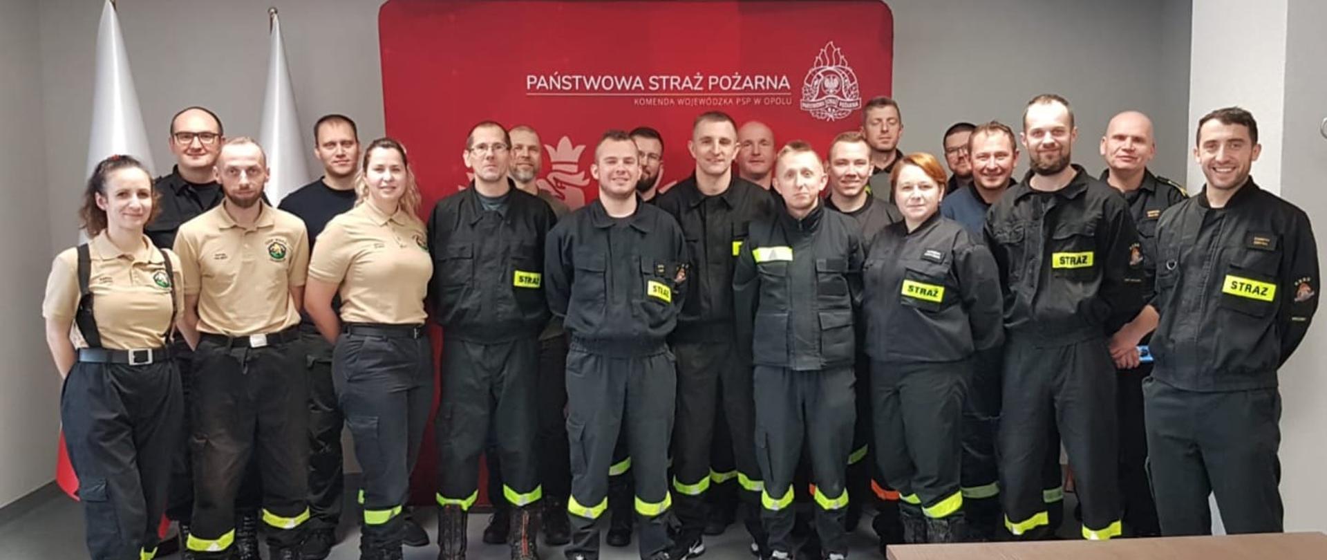 Wspólne zdjęcie grupowe strażaków po egzaminie potwierdzającym kwalifikacje ratownika kwalifikowanej pierwszej pomocy. Strażacy PSP i OSP w umundurowaniu służbowym i koszarowym stoją na tle banneru Państwowej Straży Pożarnej koloru czerwonego. Zdjęcie wykonane w sali Komendy Wojewódzkiej PSP.