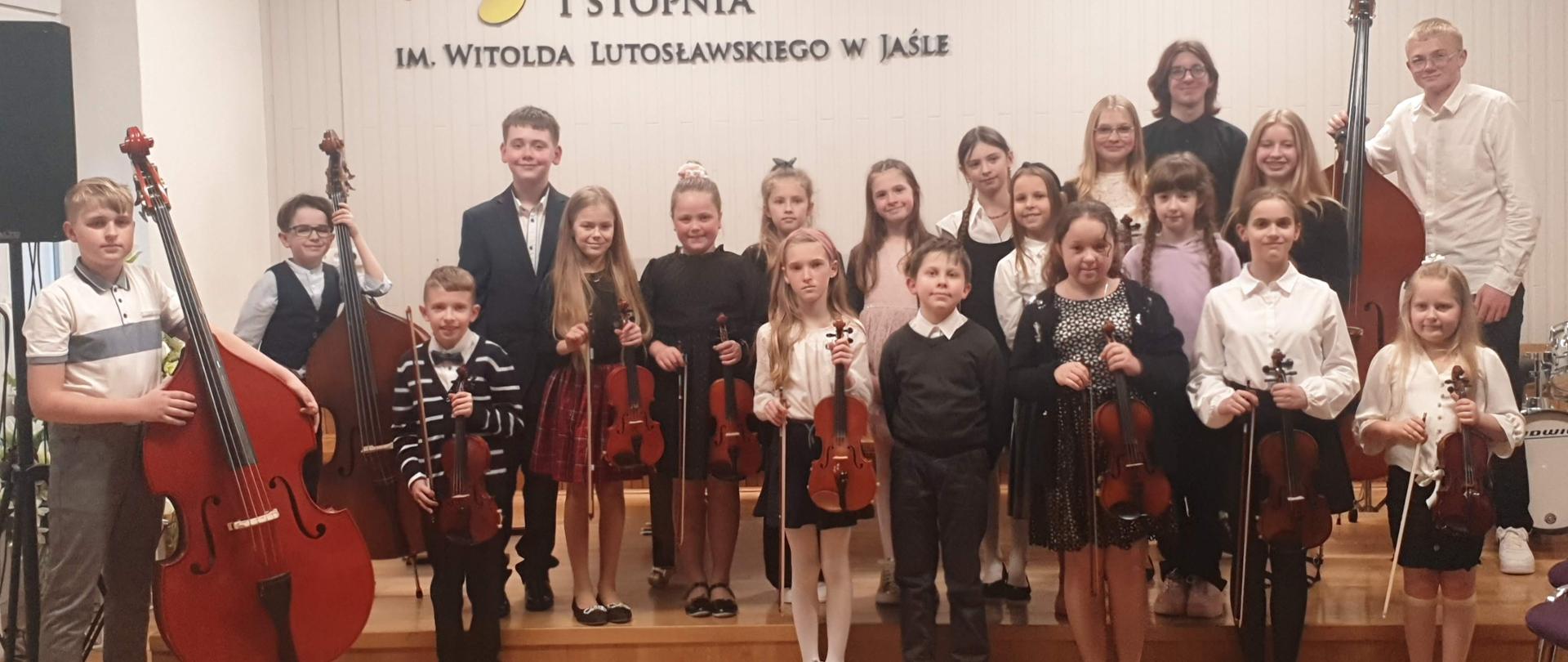 Na zdjęciu znajduje się grupa kilkunastu uczniów z instrumentami (skrzypce, kontrabasy), w tle na jasnej ścianie logo PSM oraz godło