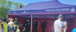 Pod granatowym namiotem strażackim z napisem Państwowa Straż Pożarna, Komenda Miejska PSP w Żorach stoją manekiny ubrane w różne stroje strażackie. przy nich oglądający ludzie. W lewej części zdjęcia widoczna część stołu. 