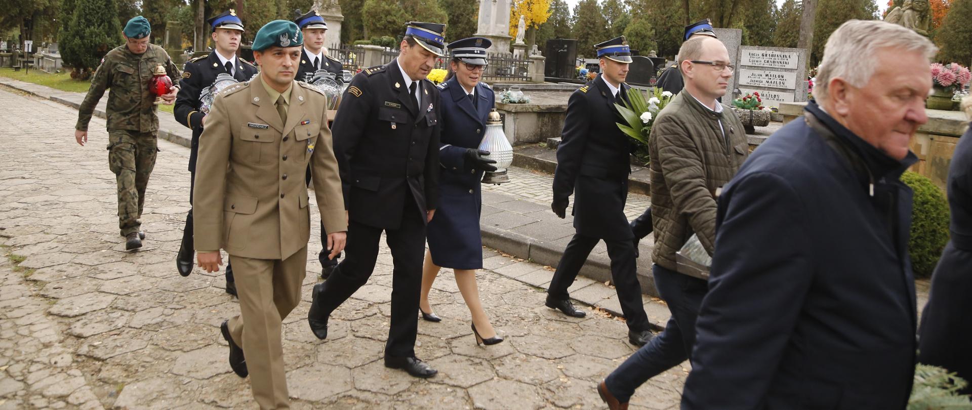 Komendant Centralnej Szkoły PSP w towarzystwie funkcjonariuszy wojska, policji, kadetów CS PSP oraz cywili kroczy aleją cmentarną