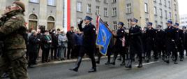 Przed budynkiem Śląskiego Urzędu Wojewódzkiego, udekorowanym biało -czerwoną wstęgą, odbywa się defilada służb mundurowych. Przed zebranymi na chodniku goścmi defiluje poczet sztandarowy oraz Kompania Reprezentacyjna Komendy Wojewódzkiej PSP w Katowicach.