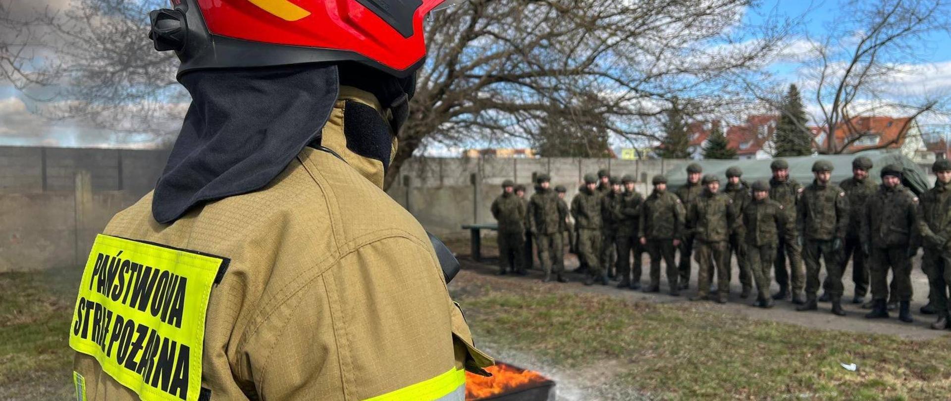 Strażak w mundurze jasnym bojowym i czerwonym hełmie. Z prawej strony grupa żołnierzy. Trwa szkolenie.
