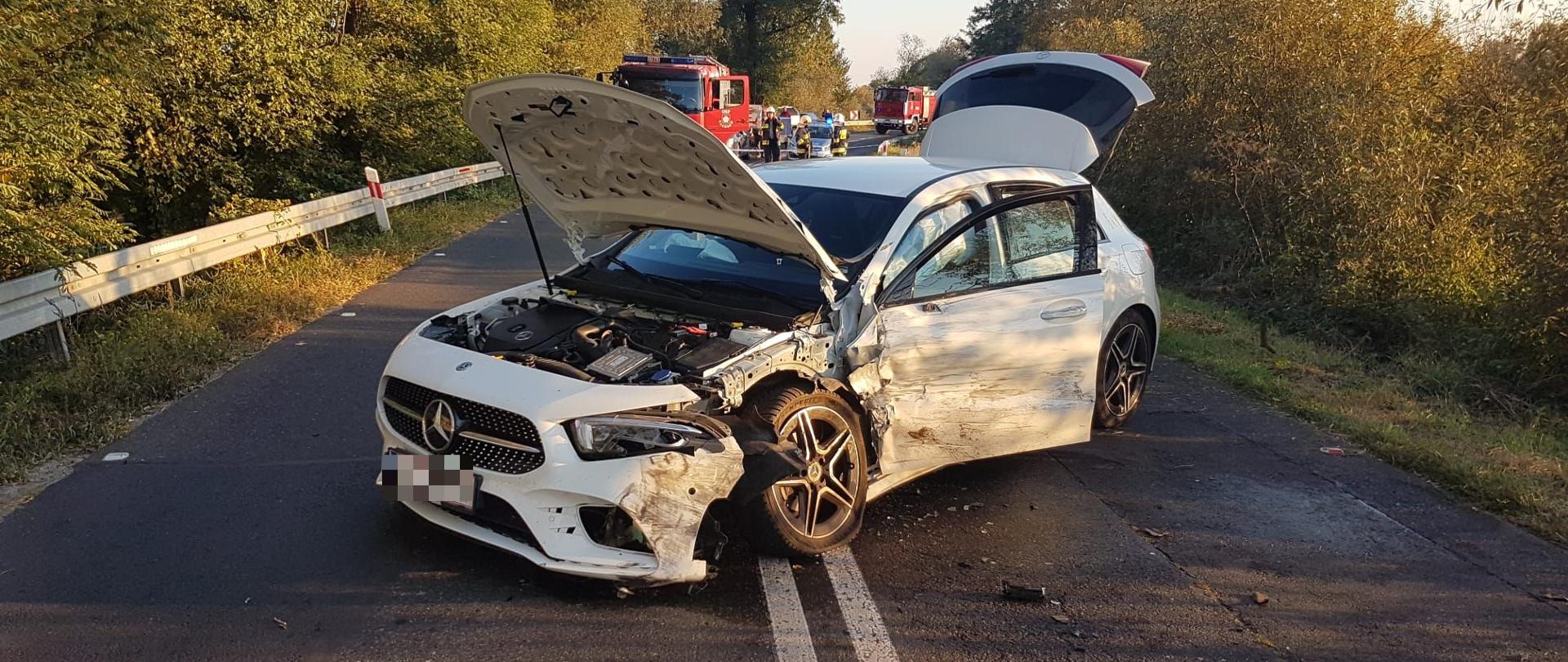 biały samochód osobowy stoi na środku drogi otwarta maska oraz klapa w oddali widać dwa samochody pożarnicze i osoby postronne, po bokach drogi widoczne drzewa oraz krzaki