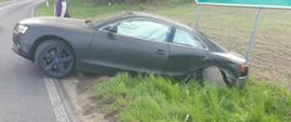Zdjęcie przedstawia stojący częściowo na poboczu i drodze czarny samochód marki Audi widoczny bokiem od strony kierowcy. Uszkodzone tylne nadkole i koło pojazdu oraz wystrzelone wewnątrz kurtyny boczne