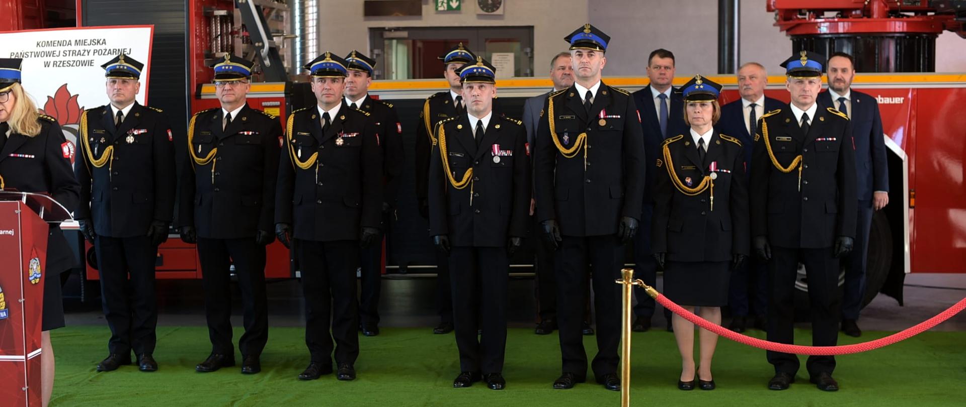 Zdjęcie przedstawia strażaków odchodzących na zaopatrzenie emerytalne oraz komendantów miejskich PSP w Rzeszowie, ubranych w mundury wyjściowe w postawie zasadniczej. Strażacy stoją na tle wozu - drabiny mechanicznej w garażu Jednostki Ratowniczo-Gaśniczej Nr 3 w Rzeszowie.