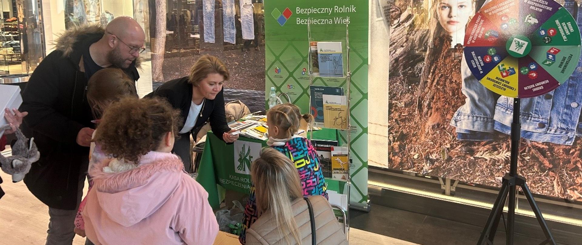 Przy stoisku KRUS w hallu galerii handlowej kobieta - pracownica KRUS - rozmawia z dziewczynką na tle rollupu KRUS i stojącego obok koła fortuny. Obok stoi mężczyzna, kobieta i dwie dziewczynki.