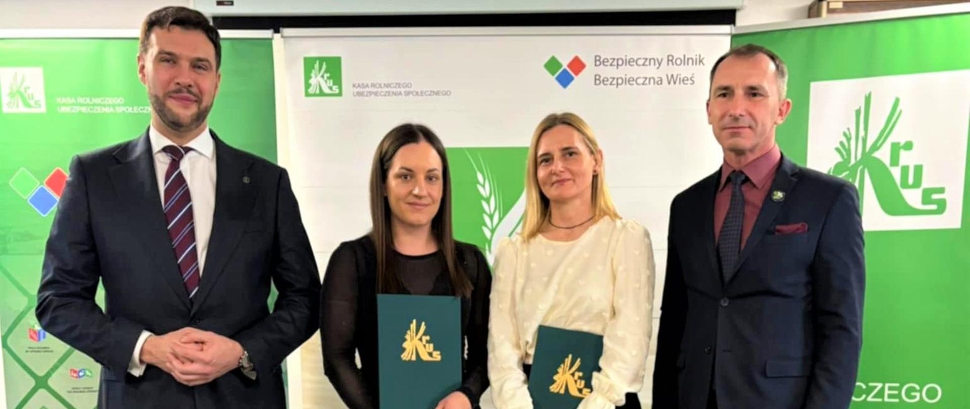 Dwie kobiety i dwóch mężczyzn pozują do zdjęcia stojąc na tle roll-upów KRUS. Obie kobiety trzymają w dłoniach zielone dyplomy ze złotym logo KRUS.