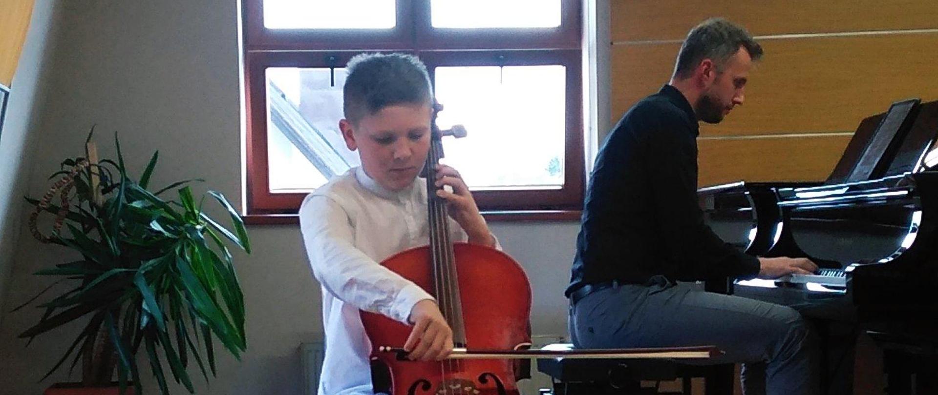 Zdjęcie na scenie sali kameralnej. Uczeń gra na wiolonczeli. Za nim z prawej strony akompaniator gra na fortepianie, następnie okno przysłonięte roletą. W rogu sali palma doniczkowa.
