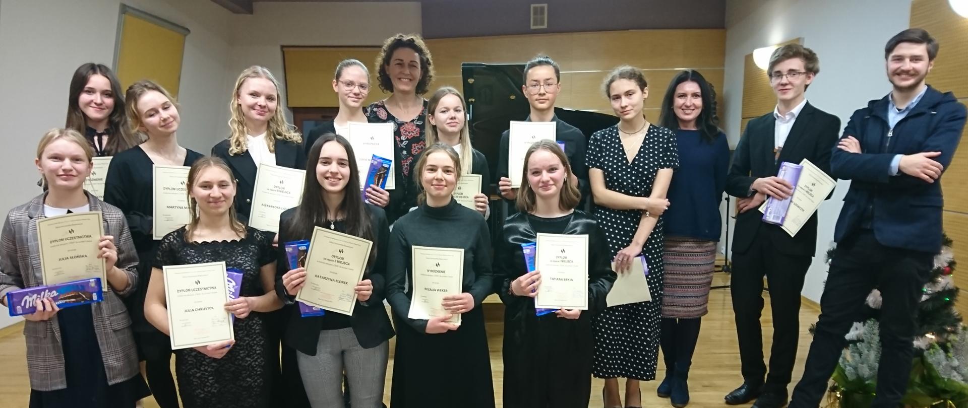 Zdjęcie przedstawia wszystkich uczestników konkursu pianistycznego Etiuda stojących w dwóch rzędach oraz ich nauczycieli. Drugi rząd na podwyższeniu. Uczestnicy trzymają dyplomy uczestnictwa, wyróżnienia oraz czekolady. 
