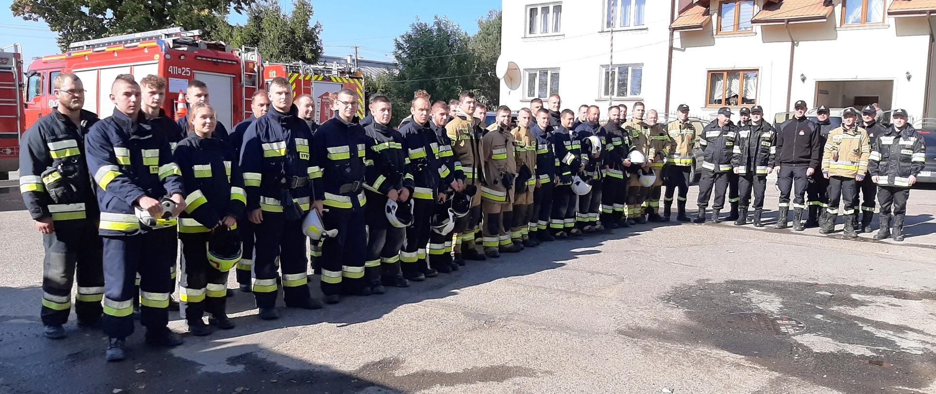 Kolorowe zdjęcie wykonane w przestrzeni otwartej w dzień. Przedstawia wielu umundurowanych strażaków na zbiórce w dwuszeregu, na utwardzonym placu, w ubraniach bojowych z hełmami w rękach. Po prawej stronie stoi kilku strażaków w ubraniach dowódczo-sztabowych. W tle widoczne czerwone samochody strażackie a w oddali widoczne częściowo budynki i drzewa.