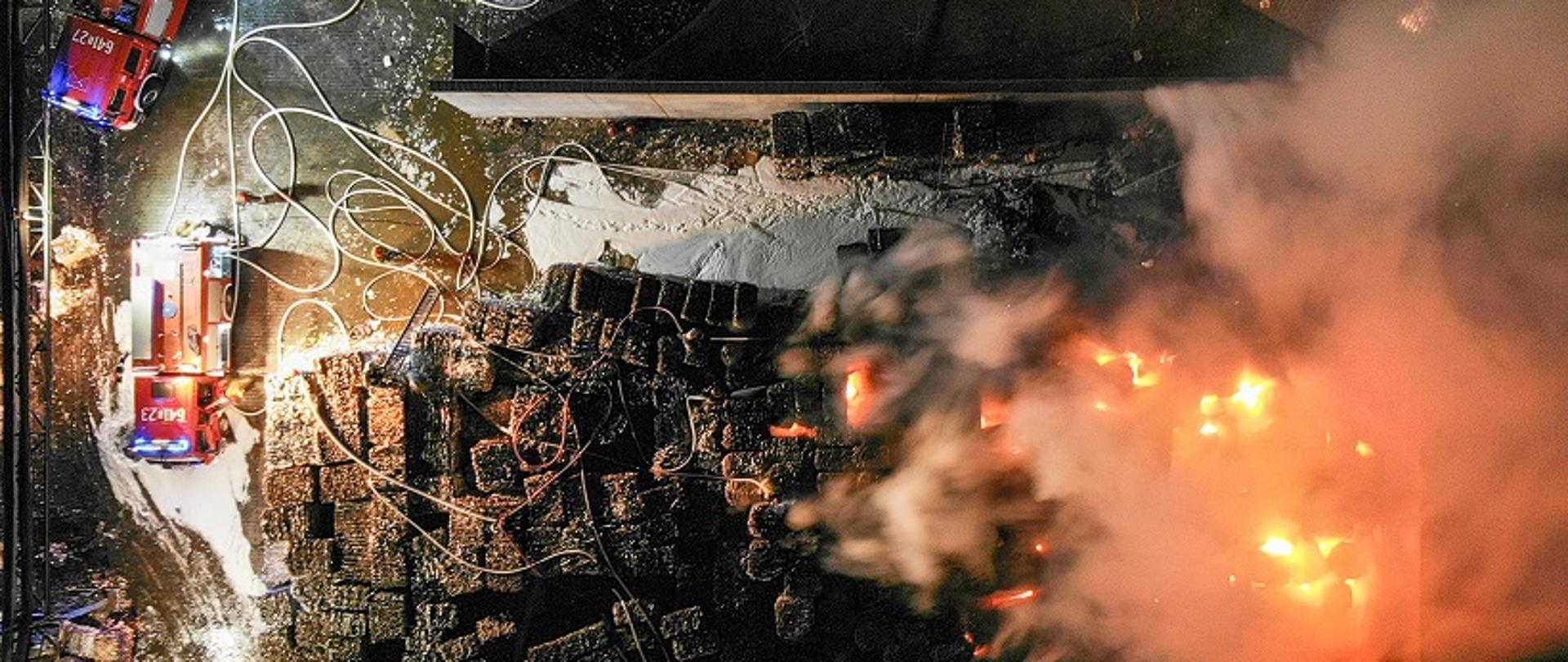 zdjęcie wykonane z drona przedstawiające widok z góry na palący się skład makulatury. Po lewej stronie zdjęcia widoczne dwa samochody ratowniczo gaśnicze z których rozwinięte są linie wężowe