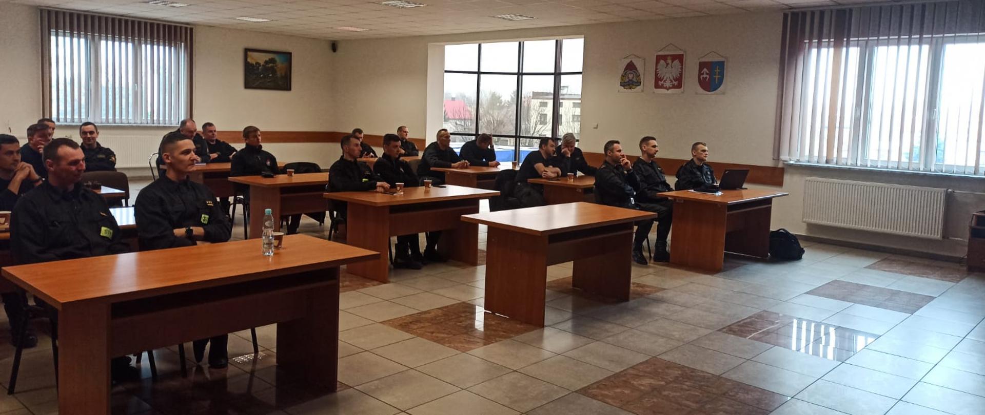 Na zdjęciu widzimy grupę strażaków podczas zajęć lekcyjnych w sali wewnątrz budynku. Mężczyźni siedzą w ławkach. Na ścianie pomiędzy oknami logo PSP, godło polski oraz znak powiatu skarżyskiego.