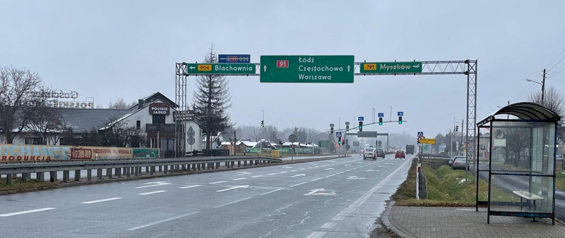 Fotografia z poziomu terenu na DK91 w Kolonii Poczesnej w rejonie skrzyżowania z ul. Kwiatową i 1 maja w kierunku Częstochowy. W prawej części fotografii znajduje się przeszklona wiata przystankowa. Środkowa cześć zajmuje 3 pasowa jezdnia rozdzielona oznakowaniem poziomy. Na horyzoncie widoczny maszt drogowej sygnalizacji świetlnej. 
