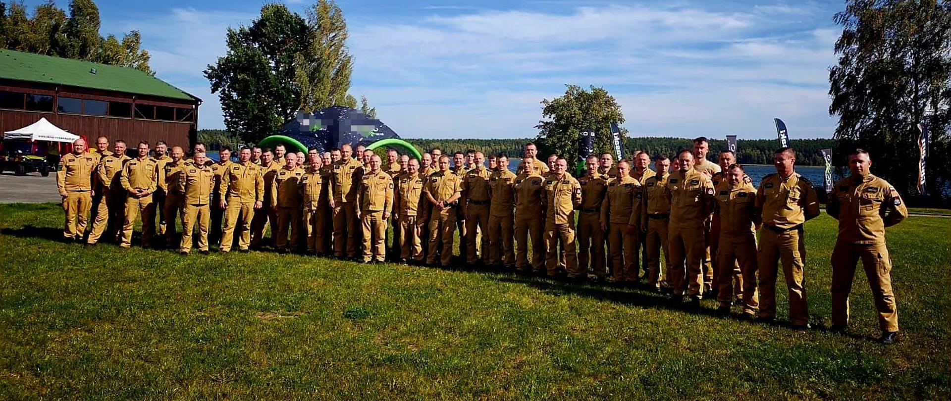 Grupa kilkudziesięciu strażaków PSP w piaskowych mundurach pozuje do wspólnego zdjęcia na trawie nad jeziorem. W tle znajdują się stoiska wystawowe i drzewa.