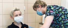 Zdjęcie przedstawia funkcjonariuszkę Państwowej Straży Pożarnej w pomieszczeniu szpitalnym punktu szczepień podczas wykonywanej iniekcji dawki szczepionki. Funkcjonariuszka siedzi na krześle szpitalnym ubrany w mundur koszarowy oraz maseczkę jednorazową. Pielęgniarka stojąca z boku dokonuje szczepienia.