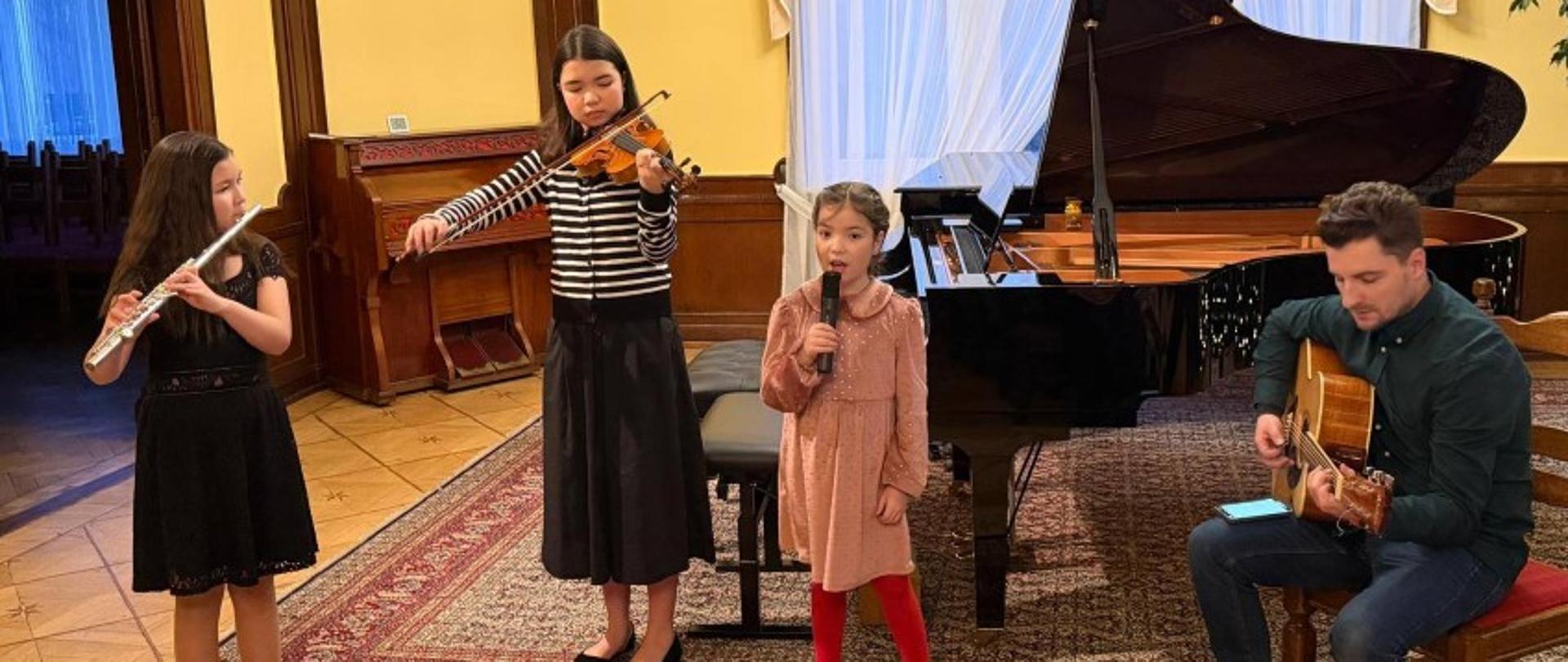 Zdjęcie kolorowe. Występ muzyczny w eleganckim wnętrzu z drewnianymi panelami i parkietem w gwiazdy. Po lewej stronie dziewczynka w czarnej sukience gra na flecie poprzecznym. Obok niej kolejna dziewczynka w pasiastej bluzce i ciemnej spódnicy gra na skrzypcach. W centrum stoi trzecia dziewczynka w różowej sukience, śpiewająca do mikrofonu. Po prawej stronie mężczyzna siedzi na krześle i gra na gitarze akustycznej. W tle widoczny jest otwarty czarny fortepian, pianino przy ścianie oraz okno z jasnymi zasłonami.