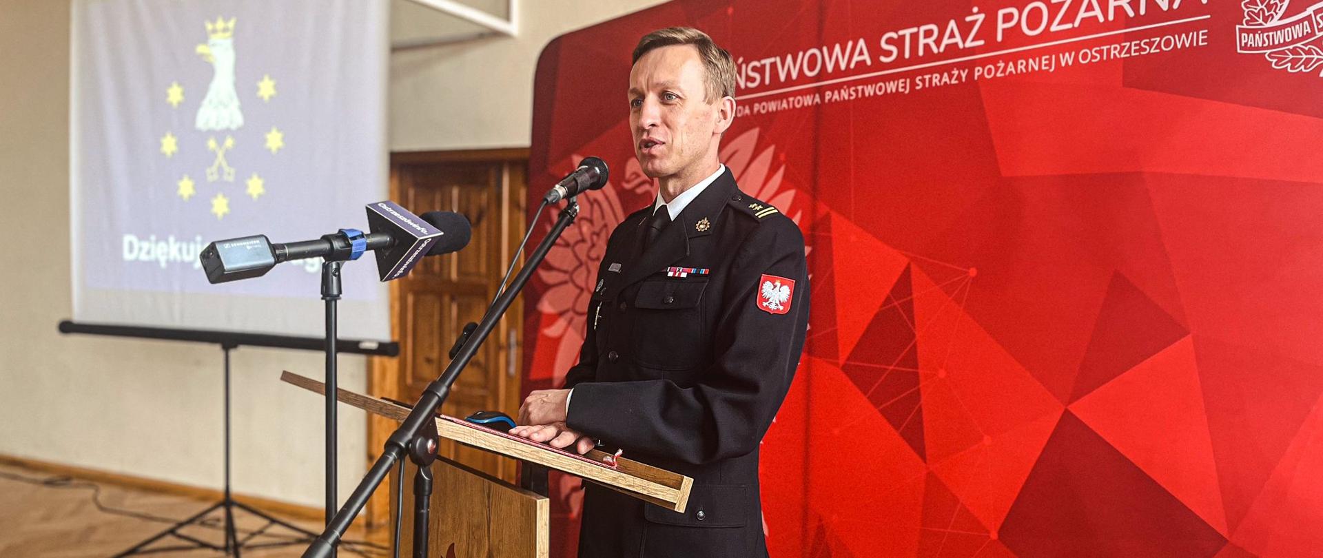 Brygadier Tomasz Grelak przemawia z mównicy w trakcie narady rocznej. W tle baner Państwowej Straży Pożarnej i ekran na którym prezentowana jest statystyka podsumowująca działania.