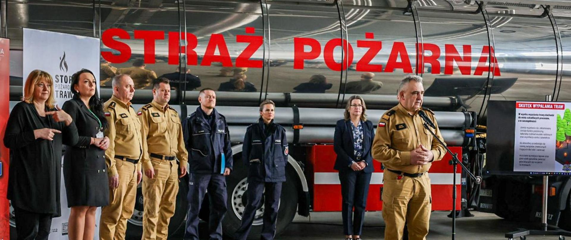 Inauguracja kampanii społecznej „Stop pożarom traw” stoją w garażu strażackim w tle srebrna cysterna strażacka