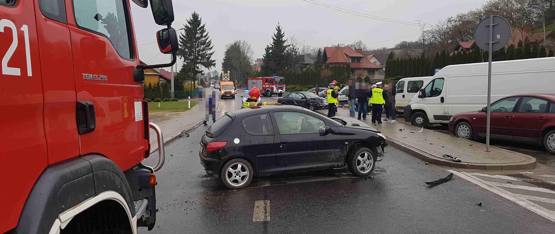 Zdjęcie przedstawia uszkodzony samochód oraz na pierwszym planie strażaka udzielając kwalifikowanej pierwszej pomocy osobie poszkodowanej. W oddali stoją pozostałe rozbite pojazdy, służba drogowa, OSP oraz Policja. 
