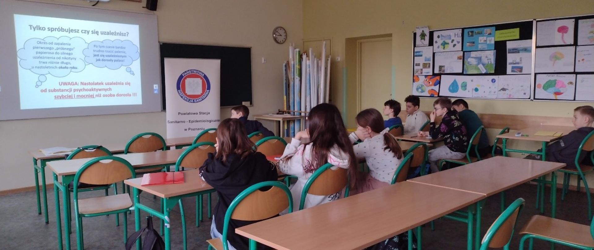 Uczniowie siedzą w ławkach i oglądają slajd z prezentacji multimedialnej zatytułowany „Tylko spróbujesz czy się uzależnisz?”