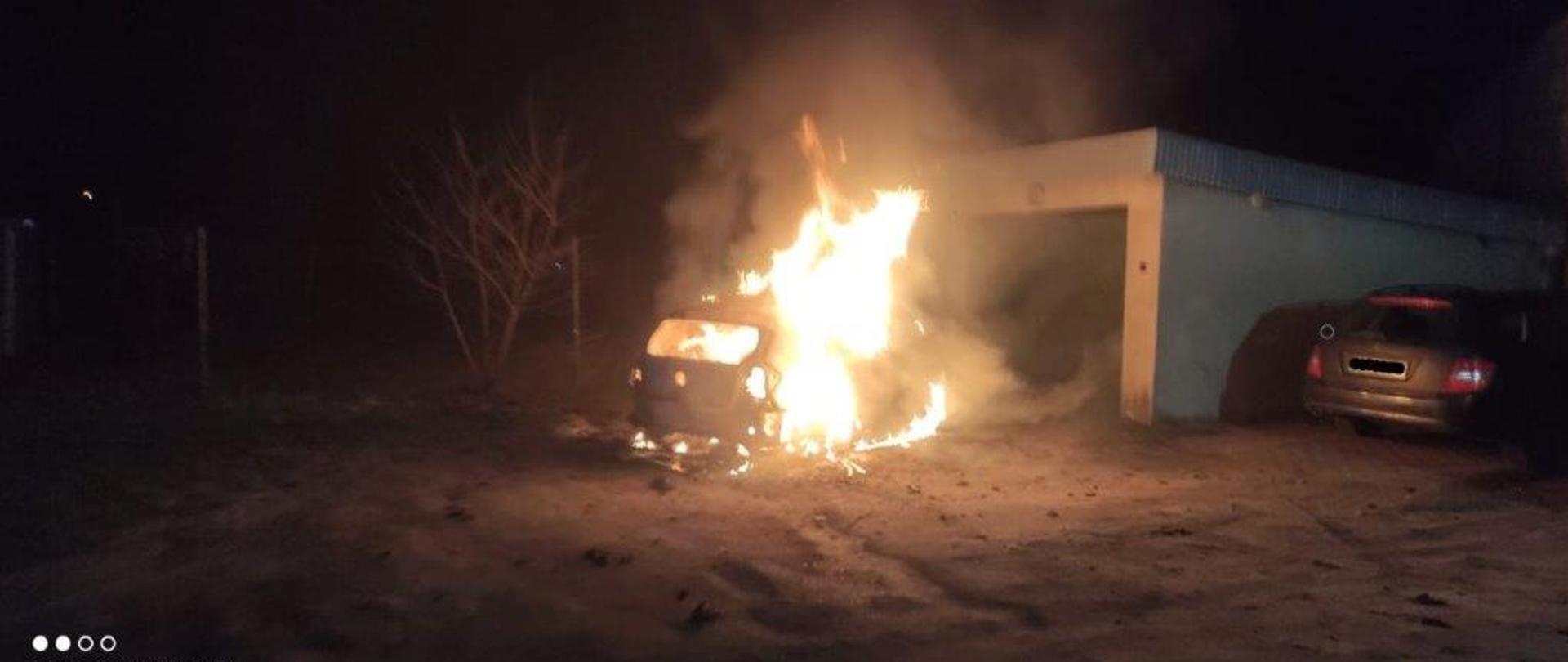 Zdjęcie przedstawia objęty pożarem samochód osobowy w porze nocnej. Samochód zaparkowany jest przy garażu.