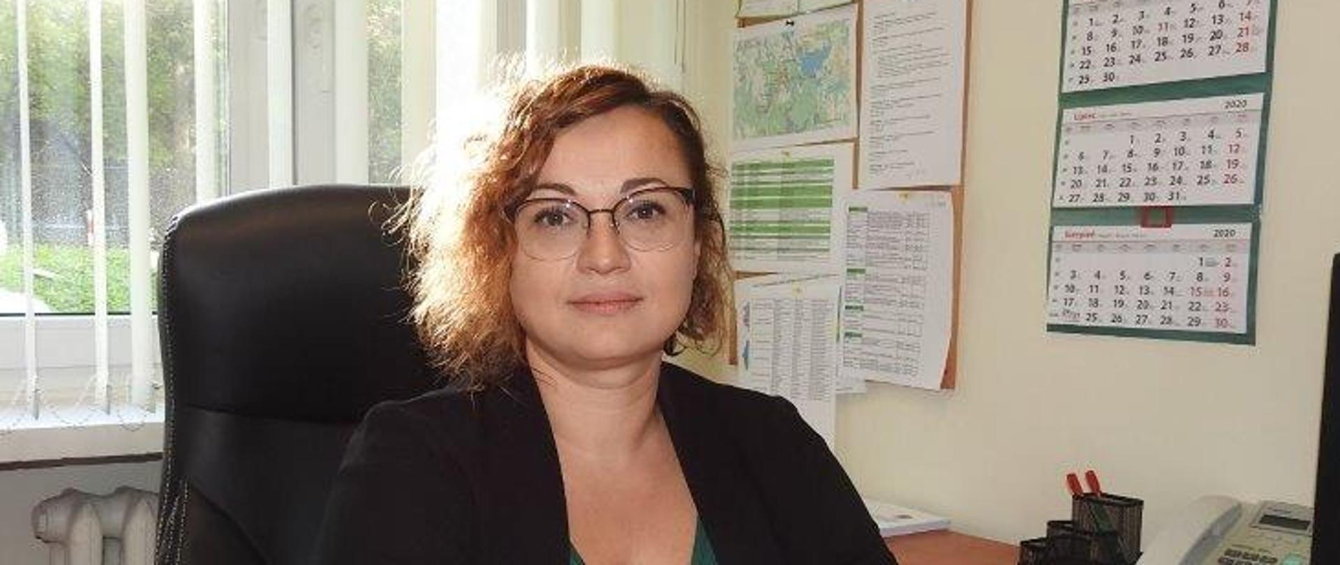 Wizerunek p.o. Zastępcy Regionalnego Dyrektora Ochrony Środowiska w Olsztynie, Regionalnego Konserwator Przyrody w Olsztynie. Kobieta siedzi przy biurku, na którym leżą dokumenty.