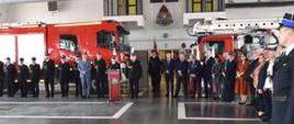 Grupa osób ubrana w umundurowanie galowe straży pożarnej lub ubrania cywilne stoi w półokręgu. W tle dwa samochody pożarnicze.