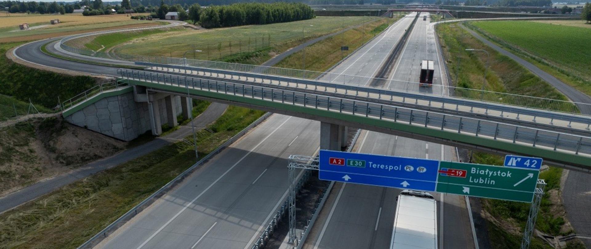 Dwujezdniowa autostrada A2 widziana z góry, z wiaduktem przebiegającym nad jezdniami i ciężarówką jadącą prawym pasem. Nad drogą znajdują się znaki kierunkowe: A2/E30 na Terespol oraz zjazd w kierunku drogi S19 na Białystok i Lublin. Wokół rozciągają się pola i tereny zielone.