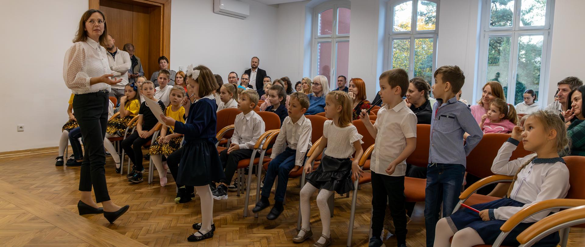 Pasowanie na ucznia. Uczniowie i publiczność zgromadzona na uroczystości podczas rozwiązywania zagadki przygotowanej przez nauczycielki.