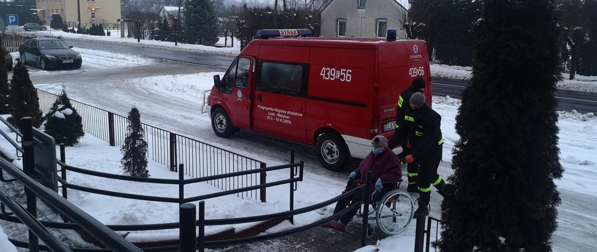 Zdjęcie przedstawia dwóch strażaków OSP pomagających osobie na wózku inwalidzkim dostać się do przychodni na szczepienie. 